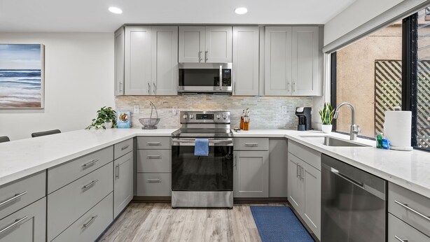 A modern kitchen with grey cabinets, stainless steel appliances, and a large window providing a view of the outdoors.