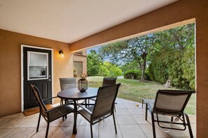 Een overdekt terras met een tafel en stoelen, een deur met een raam, en uitzicht op een tuin.