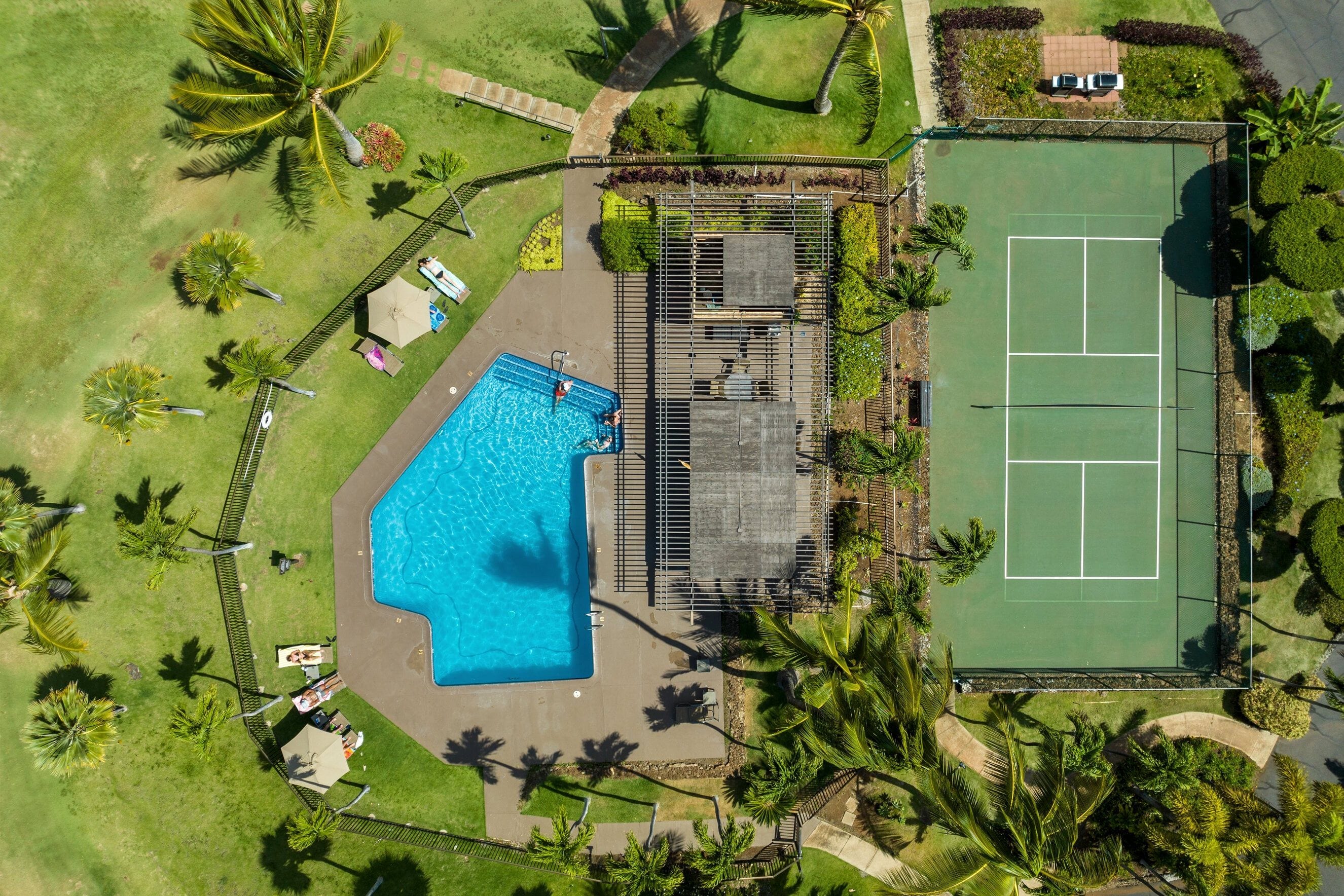 An aerial view of a property with a swimming pool, tennis court, and surrounding greenery.