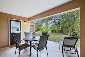 An outdoor patio with a round table and four chairs, a door with a window, and a view of a garden.