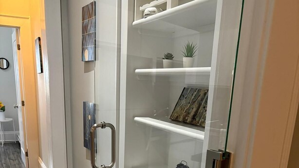 Un baño moderno con cabina de ducha de vidrio, estantes blancos con adornos y suelo de madera.