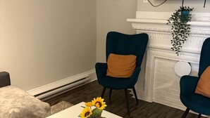 Una sala de estar con una mesa de centro blanca, dos sillones azules, un televisor montado en la pared y una chimenea.