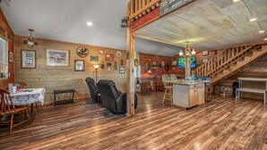 A rustic interior with wooden floors, a dining area with a table and chairs, a massage chair, a kitchen area with a sink, and a staircase leading to an upper level.