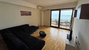 Ett rymligt vardagsrum med en svart soffa, en platt-TV och en takfläkt. En tavla hänger på väggen, och det finns en skjutdörr i glas som leder ut till en balkong med utsikt över havet.