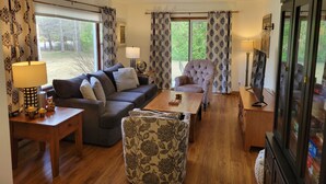 A cozy living room with a sofa, armchair, and ottoman, a wooden coffee table, and a TV. - Tranquil - North Porch Cottage - New AC - KING & QUEEN beds (Ellison Bay)
