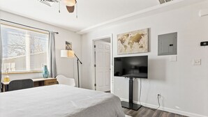 A modern hotel room with a large bed, a desk, a chair, a TV, and a window with blinds. - Central Festus Studio Apartment. Private Parking-Gigabit Wifi-Full Size Kitchen. (Festus)