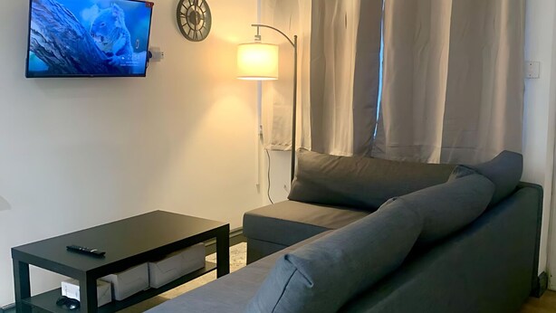 A living room with a sofa, a TV, a lamp, and a clock on the wall. - Cosy 3-Bed Home in Central Colchester (Colchester)