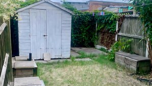 A backyard with a wooden shed, a concrete pathway, and a wooden fence covered in greenery. - Cosy 3-Bed Home in Central Colchester (Colchester)