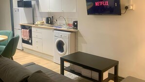 A modern kitchen with white cabinets, a washing machine, a microwave, and a sink. There's a flat-screen TV mounted on the wall displaying 'NETFLIX'. - Cosy 3-Bed Home in Central Colchester (Colchester)