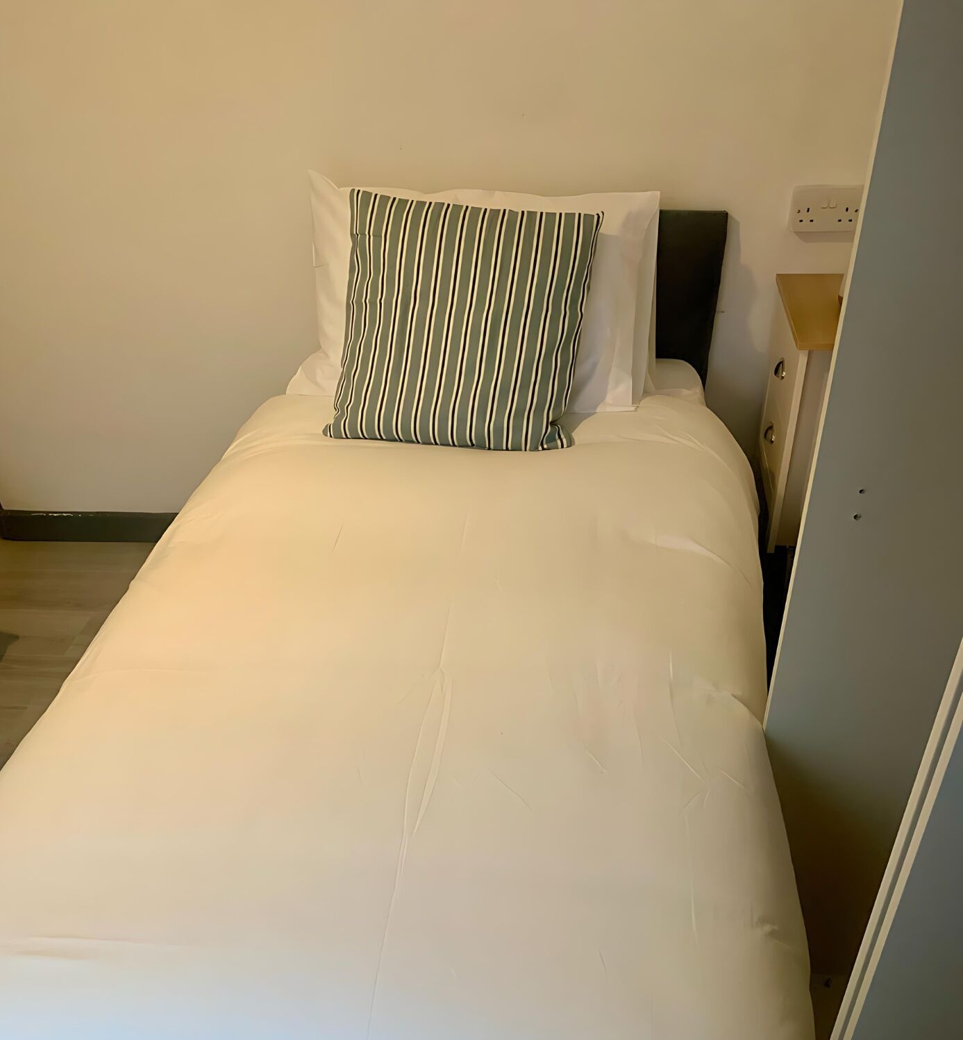 A single bed with white bedding and a striped pillow, a nightstand with a lamp, and a power outlet on the wall.