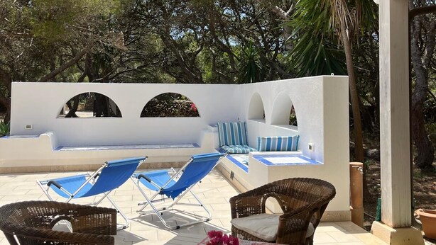 An outdoor seating area with wicker chairs and a table, a pool with blue loungers, and a white building with arches in the background.