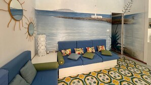 A room with a blue sofa, a patterned floor, a wall mural of a lighthouse, and a sun-shaped mirror.