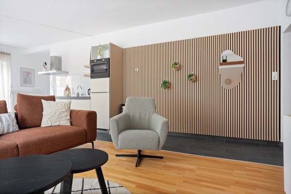 A modern living room with a sofa, armchair, and coffee table. A kitchen area with appliances and a wooden wall paneling is visible in the background.