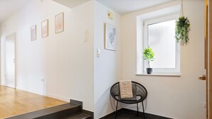 Un pasillo con piso de madera, una silla negra con una toalla, una ventana con una planta y cuadros enmarcados en la pared.