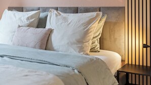 Una cama bien hecha con cabecera gris, ropa de cama blanca y una mesita de noche con lámpara.