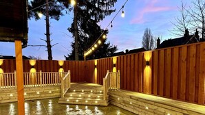 Une terrasse en bois avec un escalier menant à une zone couverte, entourée d’une clôture en bois et illuminée par des guirlandes lumineuses.