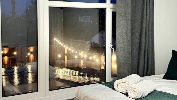 Une chambre d’hôtel dotée d’une grande fenêtre, d’un lit avec du linge de lit blanc et donnant sur un espace extérieur illuminé de guirlandes lumineuses.