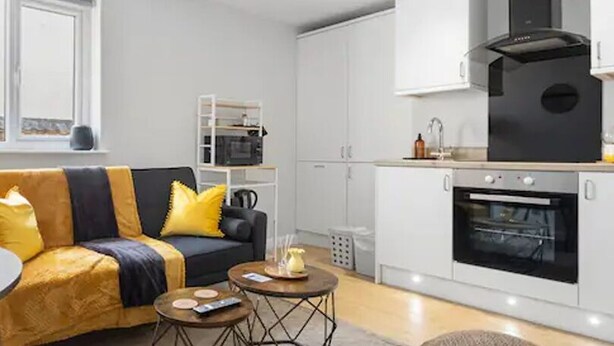 En moderne stue med sofa, sofabord og spisebord. Der er et køkkenområde med hårde hvidevarer og skabe.