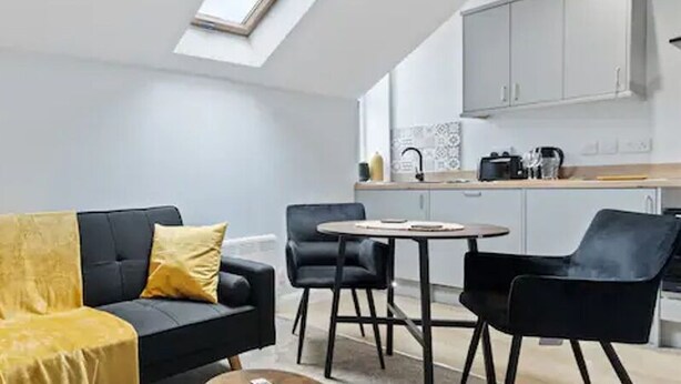 A modern living room with a black sofa, yellow cushions, a floor lamp, and a dining area with a round table and chairs. - Modern Apartment King Room |Free Parking| Suite 9 (Nottingham)