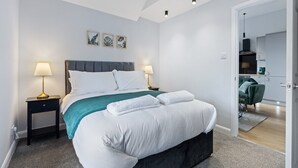 Un dormitorio moderno con una cama grande, dos mesitas de noche con lámparas, una manta verde y vistas a la cocina contigua.