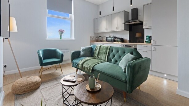 Un moderno salón con un sofá verde, un sillón azul turquesa y una mesita de centro. Se ve una ventana con vistas al cielo y una planta en el alféizar.