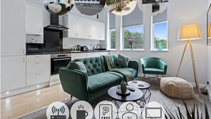 A modern living room with a green sofa, a coffee table, and a lamp. There is a kitchen area with white cabinets and a black stove. - Modern Apartment| King Bed| Free Parking |Suite 6 (Nottingham)