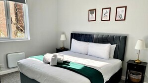 Una cama bien tendida con ropa de cama blanca y verde, un cabecero oscuro y una mesita de noche con una lámpara.
