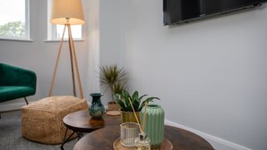 Un salon moderne avec un fauteuil vert, un lampadaire, une table basse ronde agrémentée d’objets décoratifs et un téléviseur fixé au mur.
