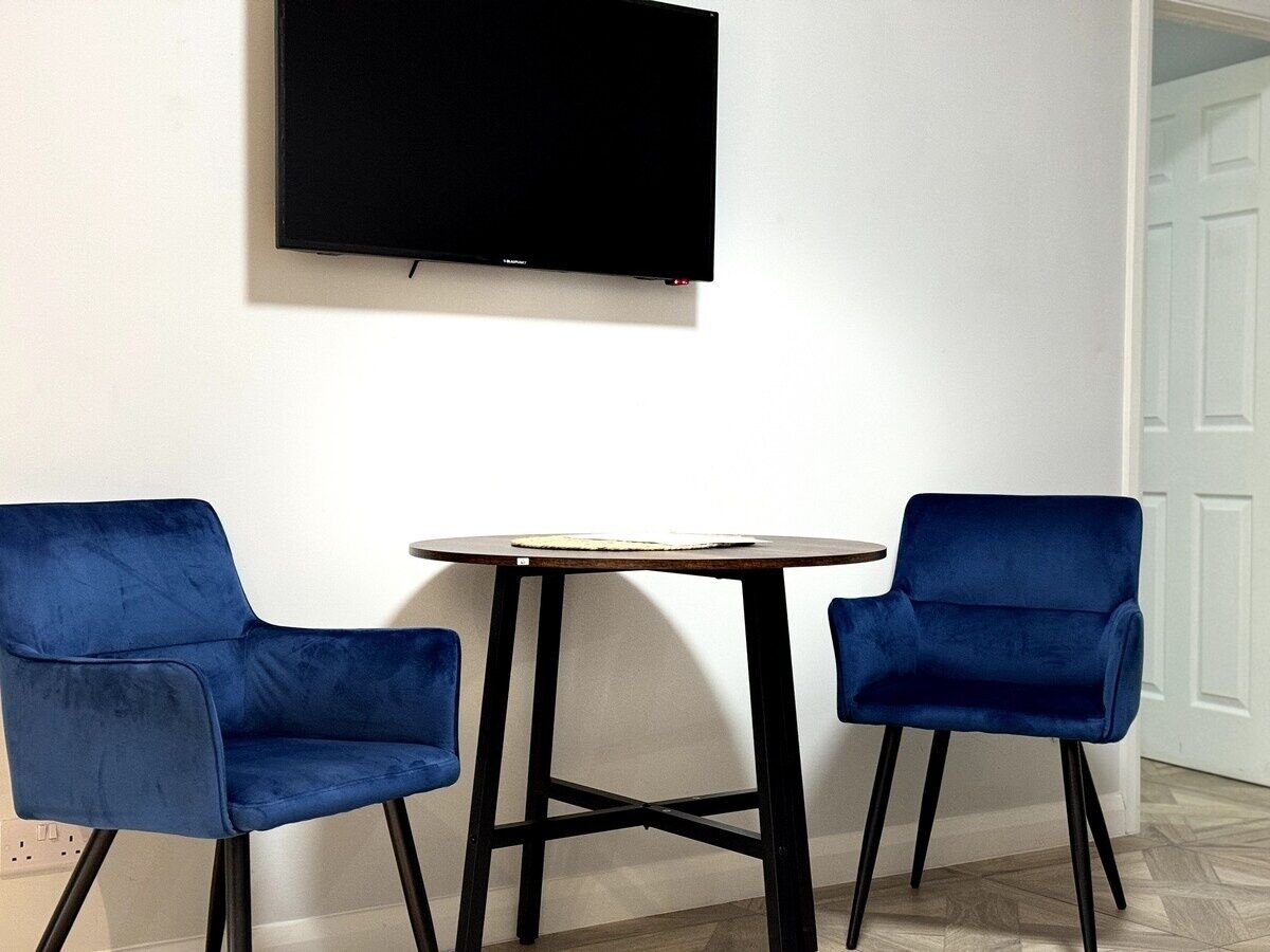 Ein Zimmer mit einem an der Wand befestigten Flachbildfernseher, einem runden Tisch mit heller Tischplatte und zwei blauen Samtstühlen.