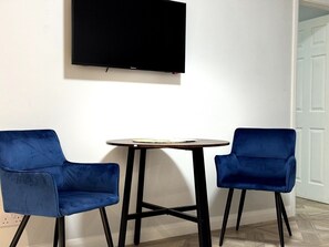 Een kamer met een wandgemonteerde flatscreen-tv, een ronde tafel met een lichtgekleurd blad en twee blauwe fluweelstoelen.