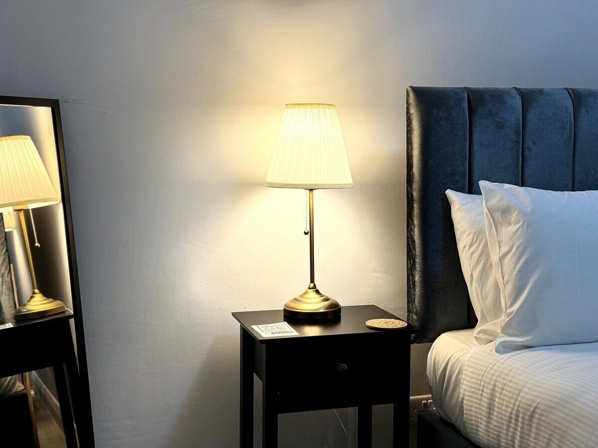 Ein Hotelzimmer mit Nachttisch, Lampe, Bett mit weißer Bettwäsche und dunklem Kopfteil.
