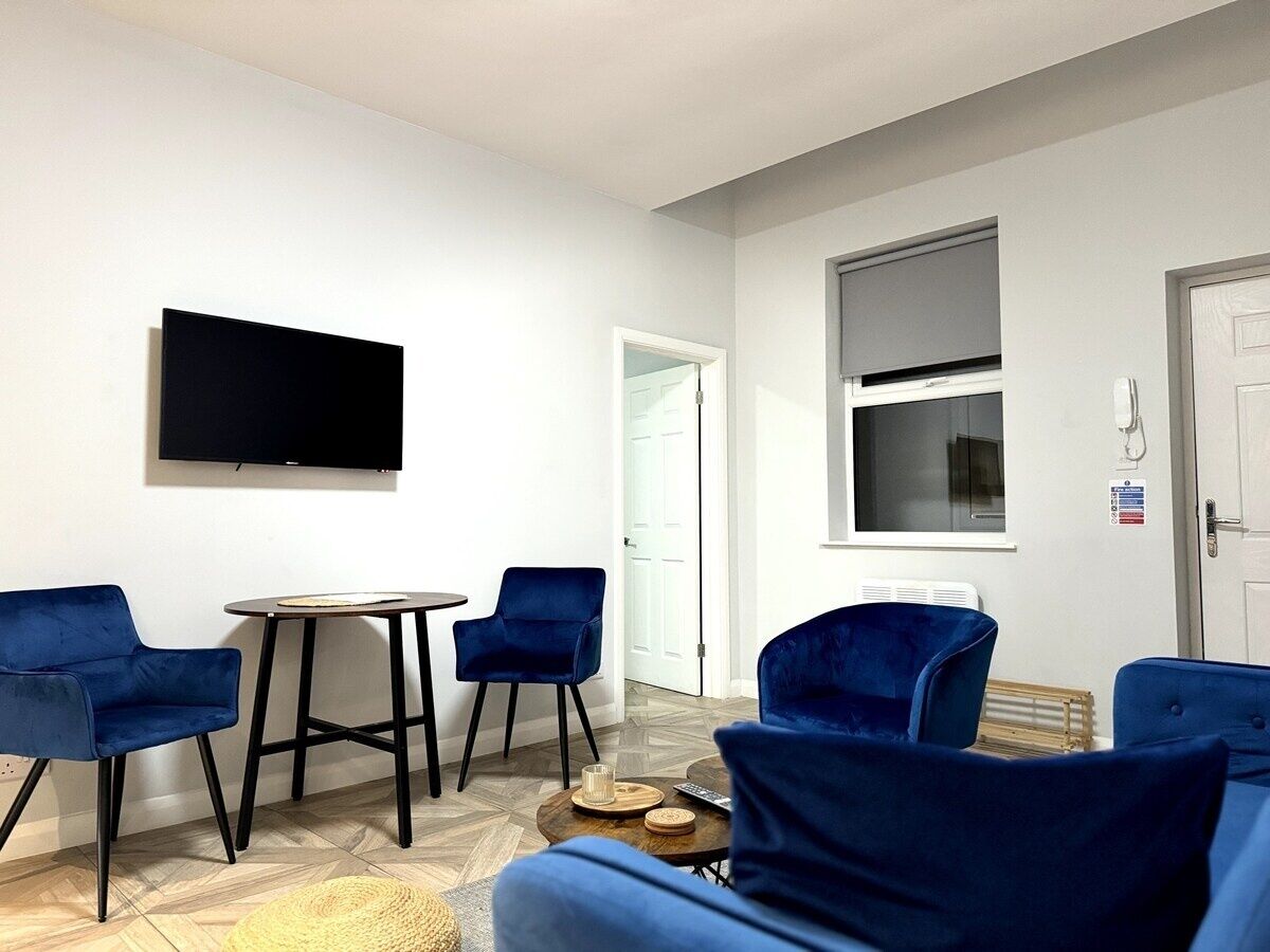 A modern living room with a flat-screen TV, two blue armchairs, a small round table, and a window with blinds.