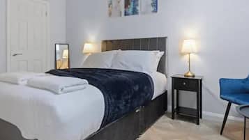 A neatly made bed with white linens and a dark blue blanket, a blue chair, a small table, and two bedside lamps.