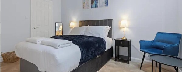A neatly made bed with white linens and a dark blue blanket, a blue chair, a small table, and two bedside lamps. - Modern Apartment Double Bed |Free Parking| Suite 4 (Nottingham)