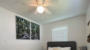 A bedroom with a bed, a ceiling fan, two wall-mounted paintings, a window with blinds, and a bedside table with a lamp. - Charming 2-bedroom Apartment near NoDa, Plaza Midwood, Uptown, and more (Charlotte)