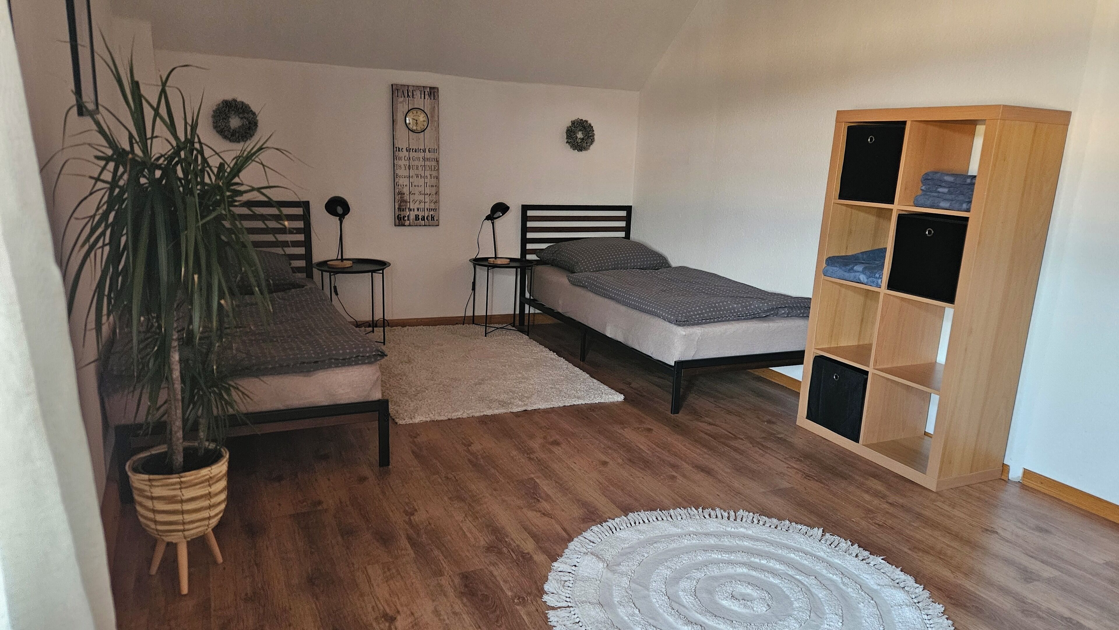 A small, cozy room with two single beds, a wooden floor, a white rug, a bookshelf, and a potted plant.