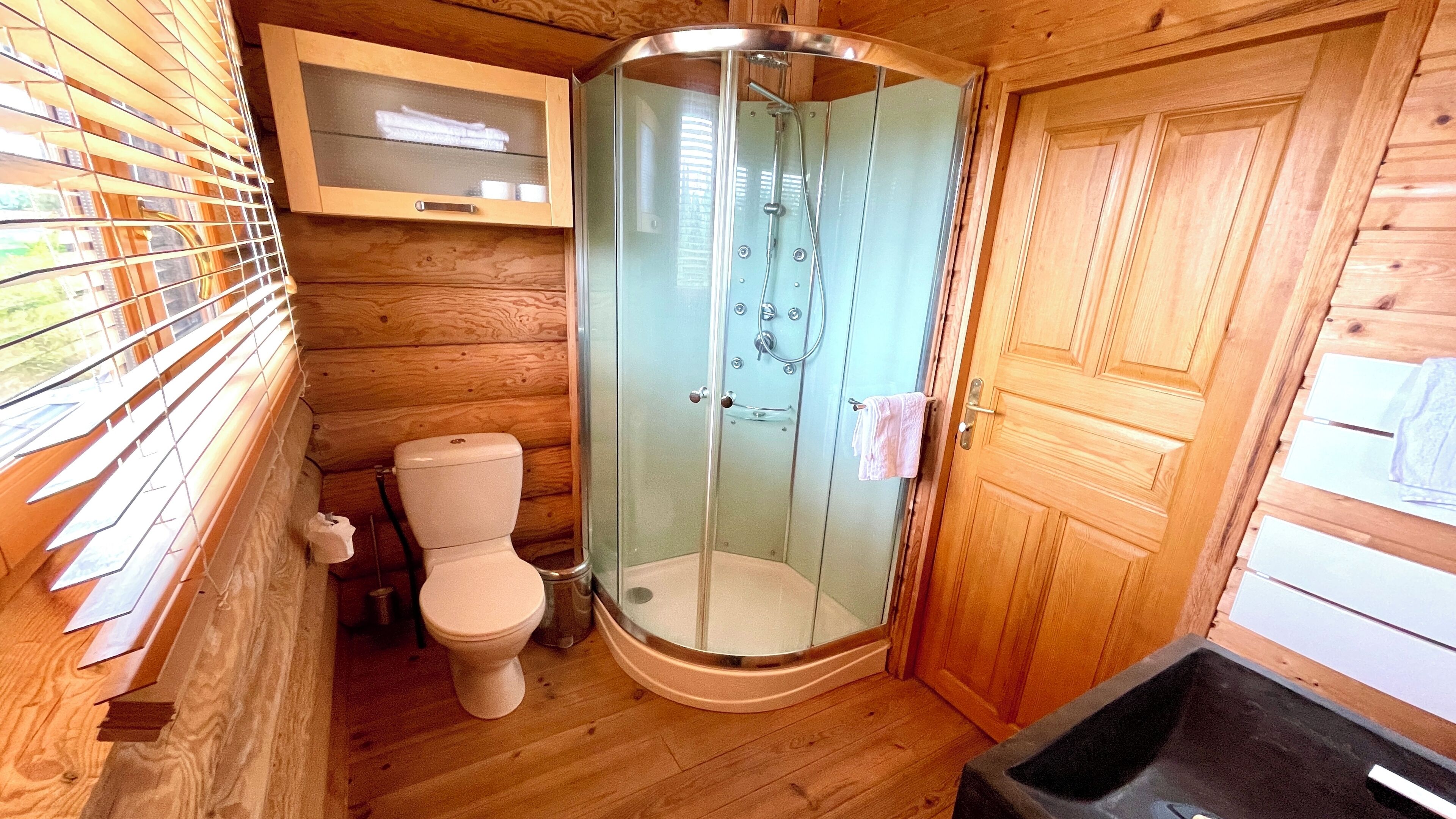 A wooden cabin bathroom with a glass shower enclosure, a toilet, and a wooden cabinet above the toilet.