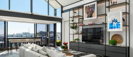 Ein modernes Wohnzimmer mit einer großen Couch, einem Fernseher und Blick auf die Stadt durch große Fenster.