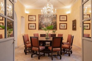 Uma sala de jantar com uma mesa redonda cercada por seis cadeiras, um candelabro e quadros nas paredes.