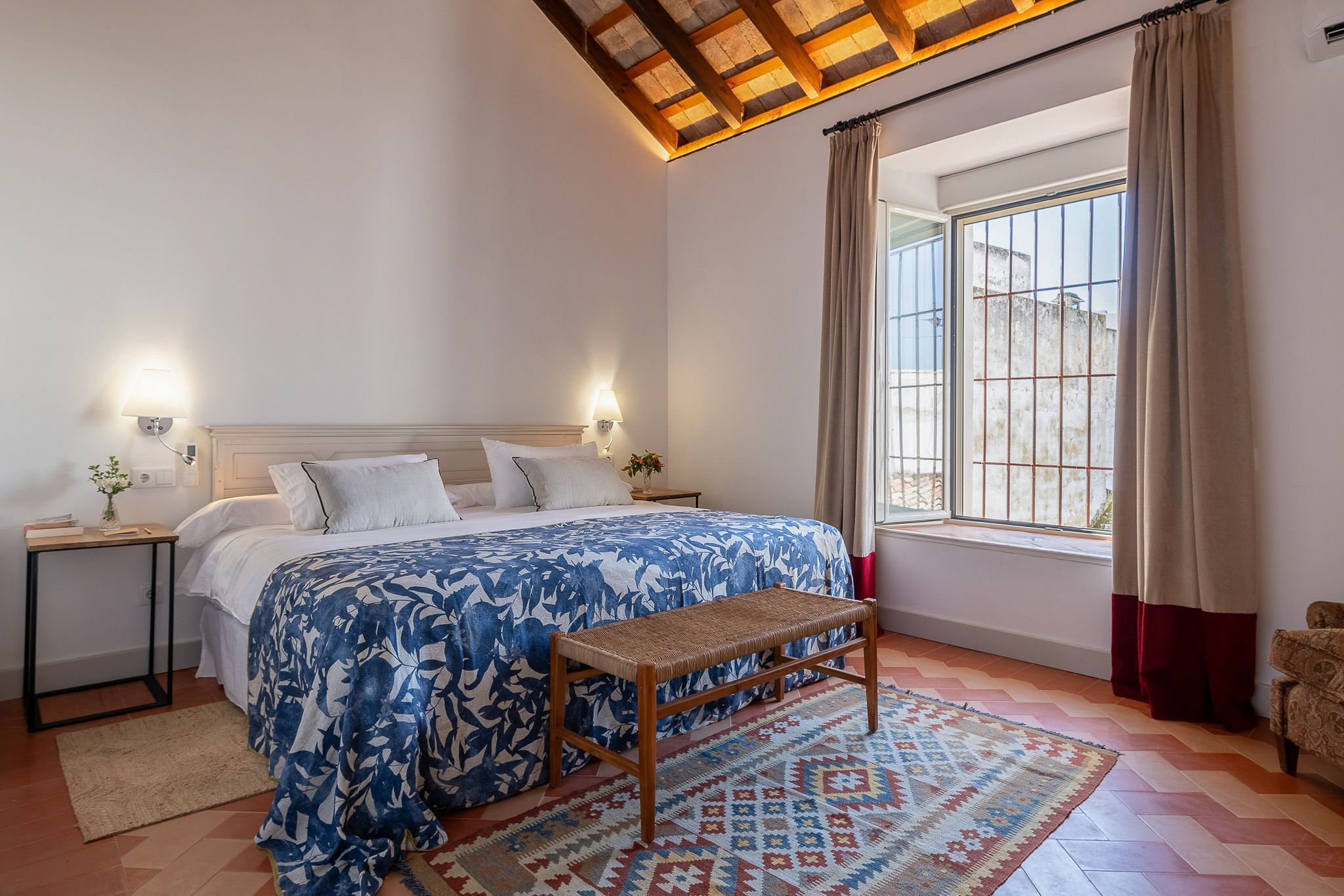 A bedroom with a bed, bedside tables, a bench, a window with curtains, and a patterned rug.