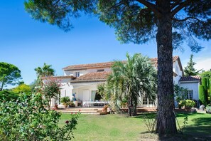 Uma casa grande com telhado telhado, rodeada por um jardim bem cuidado com palmeiras e outras plantas.