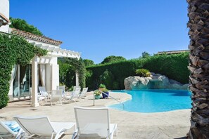 Uma área de piscina com espreguiçadeiras, um pérgola e uma formação rochosa.