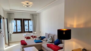 A modern living room with a sofa, a coffee table, a lamp, and a dining area with a table and chairs. - Superb Colina Smir apartment, swimming pool view in front of magnificent Kabila Beach (Marina Smir)