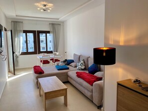 A modern living room with a sofa, a coffee table, a lamp, and a dining area with a table and chairs. - Superb Colina Smir apartment, swimming pool view in front of magnificent Kabila Beach (Marina Smir)
