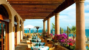 Un área de comedor al aire libre techada con mesas y sillas, columnas y vista al mar.