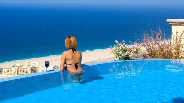 Una persona en traje de baño negro sentada en una piscina con vista al océano y a la playa.