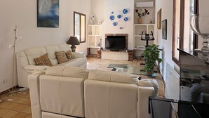 A spacious living room with a large sofa, a flat-screen TV, and a coffee table. There is a fireplace, a bookshelf, and a window with curtains. - Family home with pool (Saint-Didier)