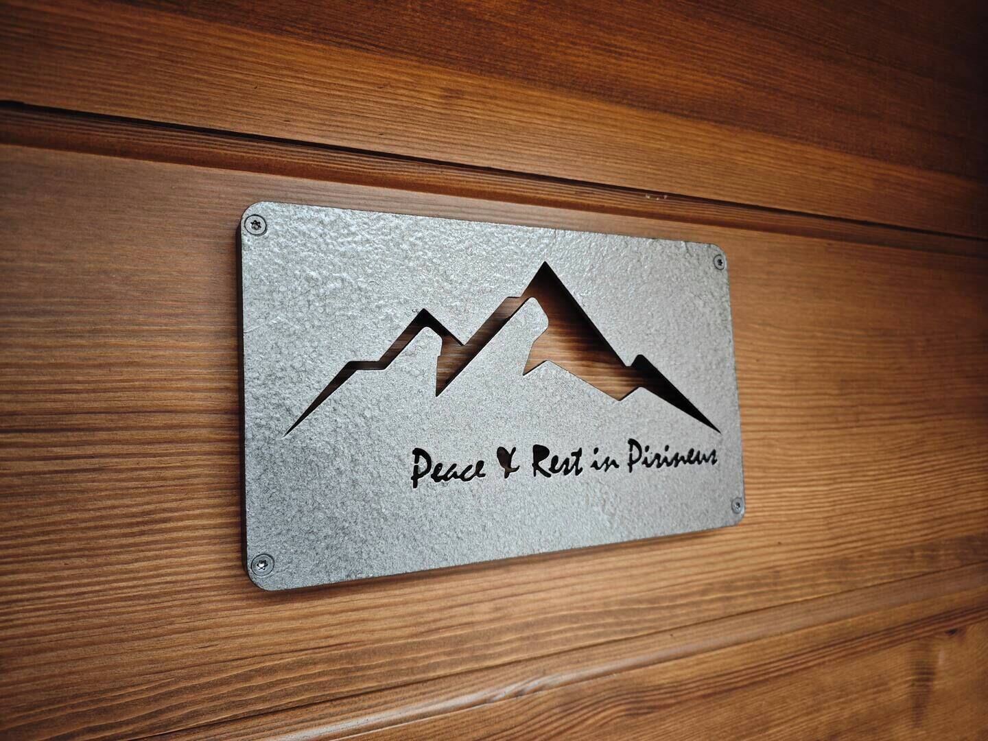 Una placa de metal con un diseño de montaña y el texto 'Peace & Rest in Pirineum' sobre una superficie de madera.