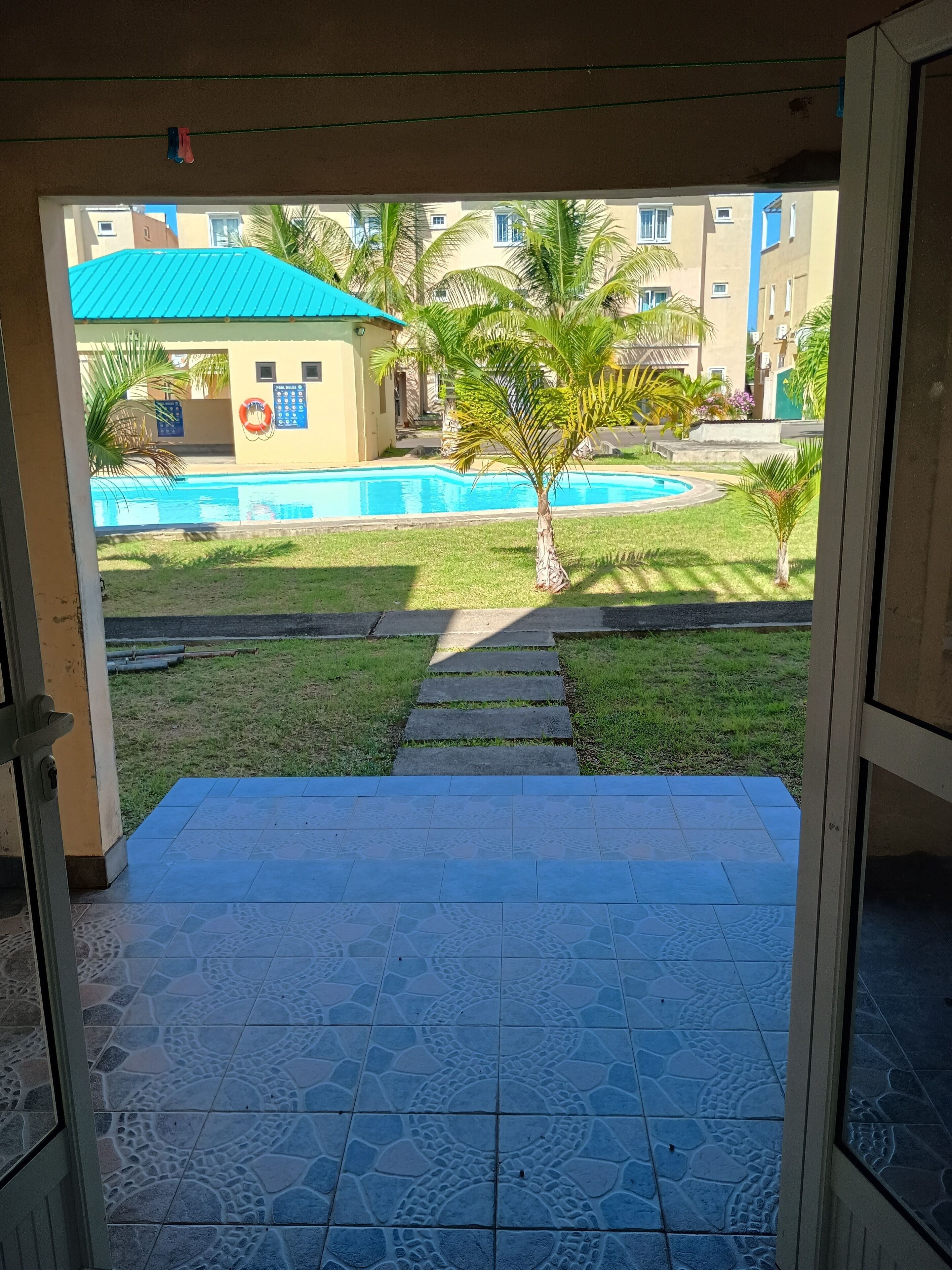A view from a room to a swimming pool area with a tiled floor, palm trees, and a building with a blue roof.