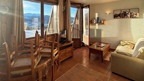 Un salón espacioso con una mesa de comedor de madera, sillas, un sofá y una televisión. Amplias ventanas ofrecen vistas a montañas nevadas.